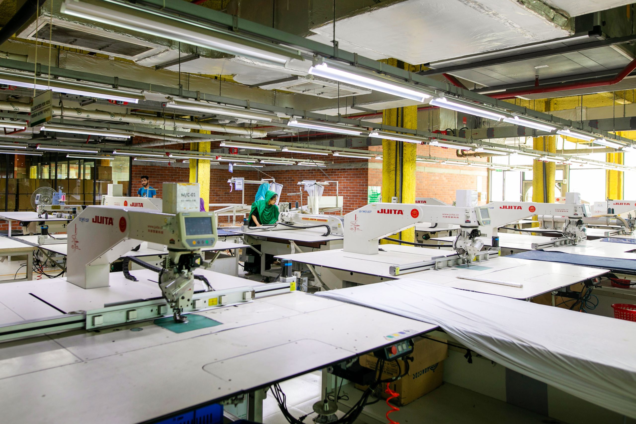 a single factory worker in the midst of many workstation tables under industrial lighting fixtures