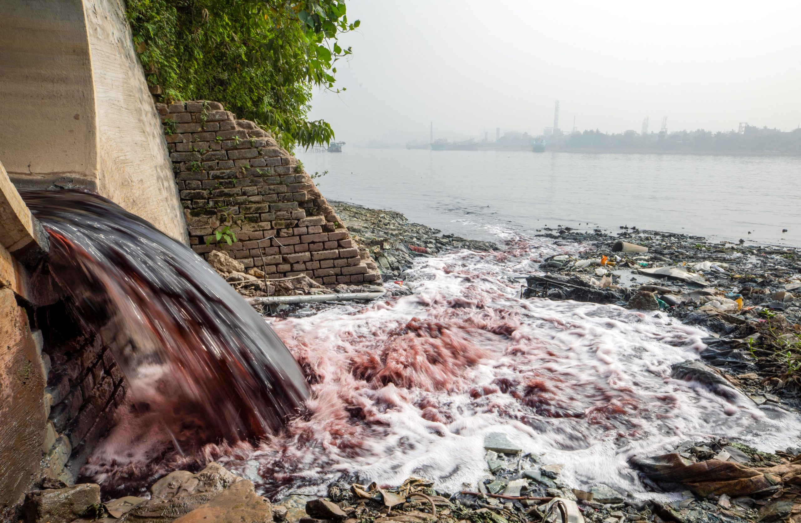 colored water pouring out of a cement tunnel into a river with a city in the far distance